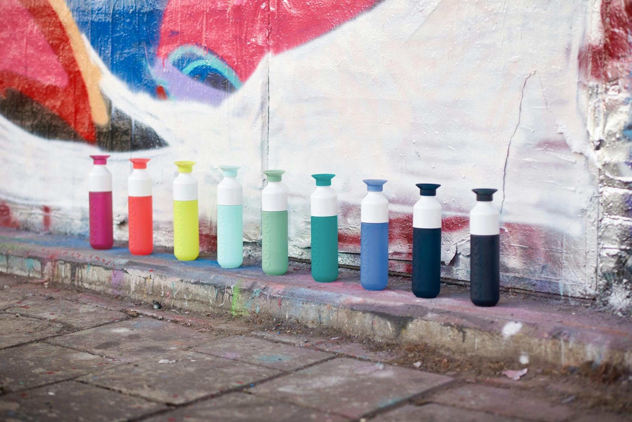 Line up of 9 colourful Dopper Original bottles in front of graffiti